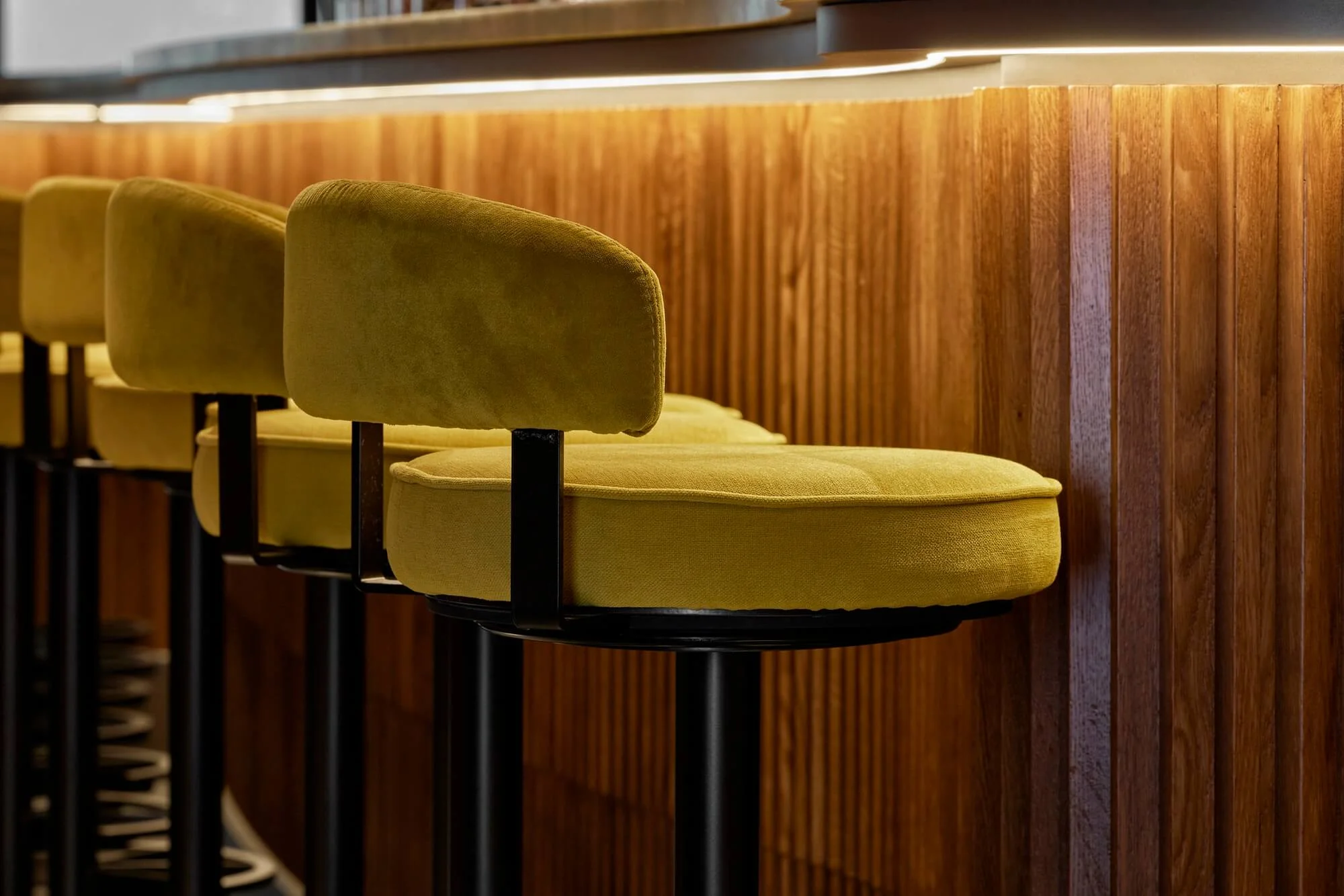 A low-angle close-up of a stool's back and black metal legs, with the fluted bar counter backlit behind it